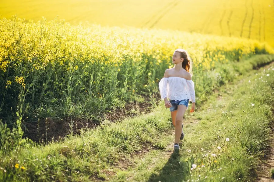 A young woman walking joyfully through a sunlit field of yellow flowers, representing the return to vitality and health through natural autoimmune support.